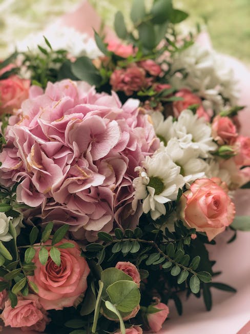 A close-up view of a floral bouquet featuring large, soft pink hydrangea blooms with ruffled petals, complemented by delicate pale pink roses and white daisies with yellow centers. The arrangement inc