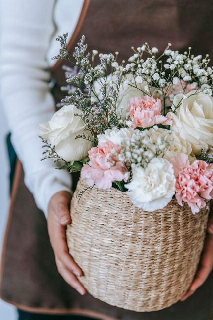 A person holding a round, woven wicker basket filled with an arrangement of fresh flowers, including white roses, soft pink carnations, sprigs of delicate gypsophila, and purple lavender. The flowers 