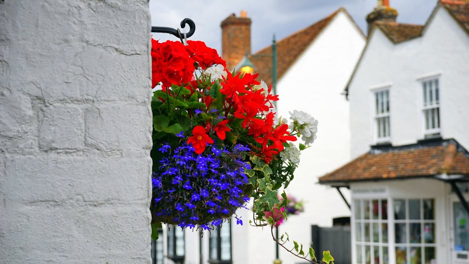 A vibrant hanging flower basket attached to a white brick wall, featuring a mix of red geraniums, white petunias, and blue lobelias, with green foliage interspersed. The basket is suspended by a black