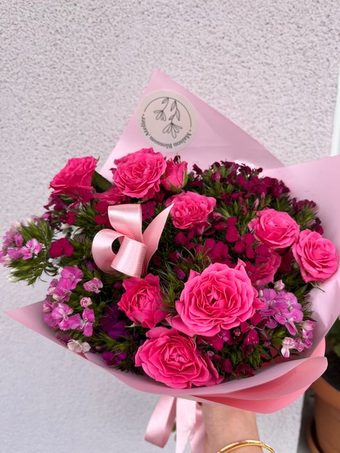 A vibrant bouquet of fresh pink roses, complemented by smaller purple and pink flowers, arranged with lush green foliage. The flowers are wrapped in light pink tissue paper and secured with a satin pi