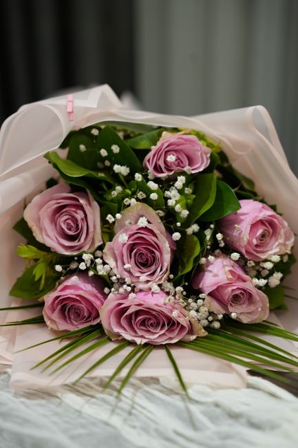 A bouquet of fresh pink roses with full, velvety petals arranged in a circular pattern, complemented by small white baby's breath flowers and lush green foliage. The bouquet is wrapped in elegant whit