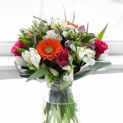 Gracefully arranged and awash in harmonious hues, this bouquet channels the essence of nature's elegance found throughout Beckenham's leafy boulevards and tranquil gardens. The clear glass vase reveals a tangle of fresh, green stems, underscoring authenticity and craftsmanship. Large gerbera daisies-two in a spirited orange, two in a creamy peach-anchor the design with sun-kissed vibrance and tranquil warmth. They frame lush magenta roses, their tightly coiled petals adding rich layers, drama, and a soft, alluring fragrance to the overall composition. Dotted throughout are clusters of white alstroemeria and minute spray roses, infusing the bouquet with gentle highlights of purity and serving as a delicate counterpoint to the more opulent blooms. Light pink petals and a cascade of airy, tiny white flowers add an understated charm-a subtle nod to the romantic alleys winding off Beckenham High Street. Enveloping all, glossy green foliage offers contrast and structural integrity, while wide, paddle-shaped aspidistra leaves create a protective mantel, elegantly cupped around the bouquet's perimeter. Smaller pointed green leaves and wisps of assorted greenery fill the gaps, evoking a wild, garden-fresh beauty. Bathed in natural daylight, every colour and texture is heightened; the atmosphere feels rejuvenating and serene, apt for new beginnings, milestone celebrations, or moments requiring an expression of sincere gratitude. The arrangement's confident, yet intimate aura makes it perfect for gifting, enhancing a home along Bromley Road, or marking special occasions within the Beckenham community.